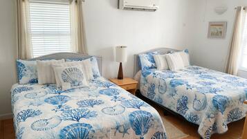 A bedroom with two beds, each with a blue and white floral bedspread, a bedside table with a lamp, and an air conditioning unit on the wall.