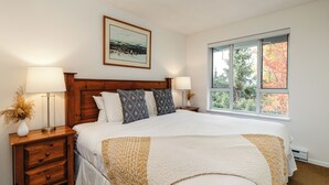 A bedroom with a large bed, two bedside tables with lamps, a framed picture on the wall, and a window with a view of trees. - Whistler Village Stroll Condo with AC and Free Parking (Whistler)