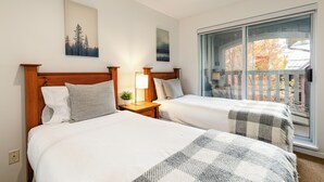 A hotel room with two beds, a wooden headboard, a nightstand with a lamp, and a view of autumn trees through a large window. - Whistler Village Stroll Condo with AC and Free Parking (Whistler)