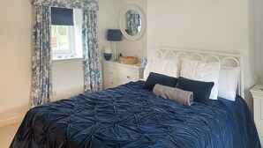 Een slaapkamer met een groot bed, een nachtkastje, een dressoir, een raam met gordijnen, een lamp en een spiegel.