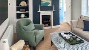 Een woonkamer met een open haard, een groene fauteuil, een gestreept voetenbankje en een boekenkast.