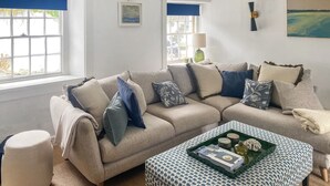 Een woonkamer met een beige bank, een gestreept voetenbankje en een raam met blauwe gordijnen.