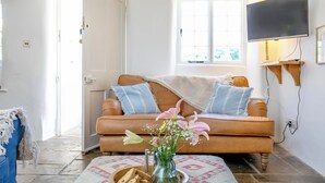 A cozy living room with a brown leather sofa, a wooden coffee table with a vase of flowers and cookies, and a flat-screen TV mounted on the wall. - 4 bedroom accommodation in Portscatho (Portscatho)