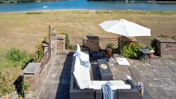 An outdoor patio with a table and chairs, a white umbrella, and a view of a lake and hills. - 4 bedroom accommodation in Portscatho (Portscatho)