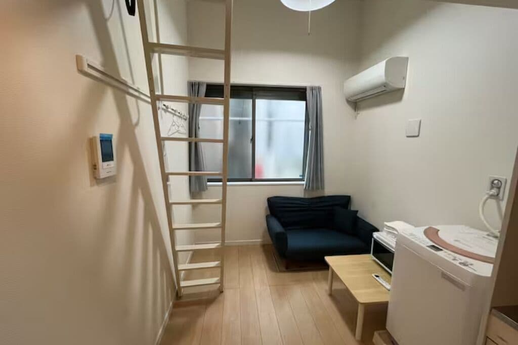 A small room with a wooden ladder, a dark blue sofa, a small table, and a compact washer-dryer unit.