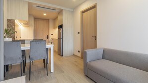 Uma sala de estar moderna com um sofá cinzento, uma área de jantar com uma mesa branca e cadeiras cinzas, e uma cozinha com armários de madeira e forno embutido.