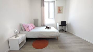 A bedroom with a bed, a desk, a chair, a radiator, a window with curtains, and a painting on the wall.