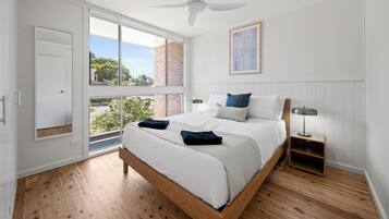 Quarto com uma grande janela, ventilador de teto, cama com almofadas, mesa de cabeceira e quadro na parede.