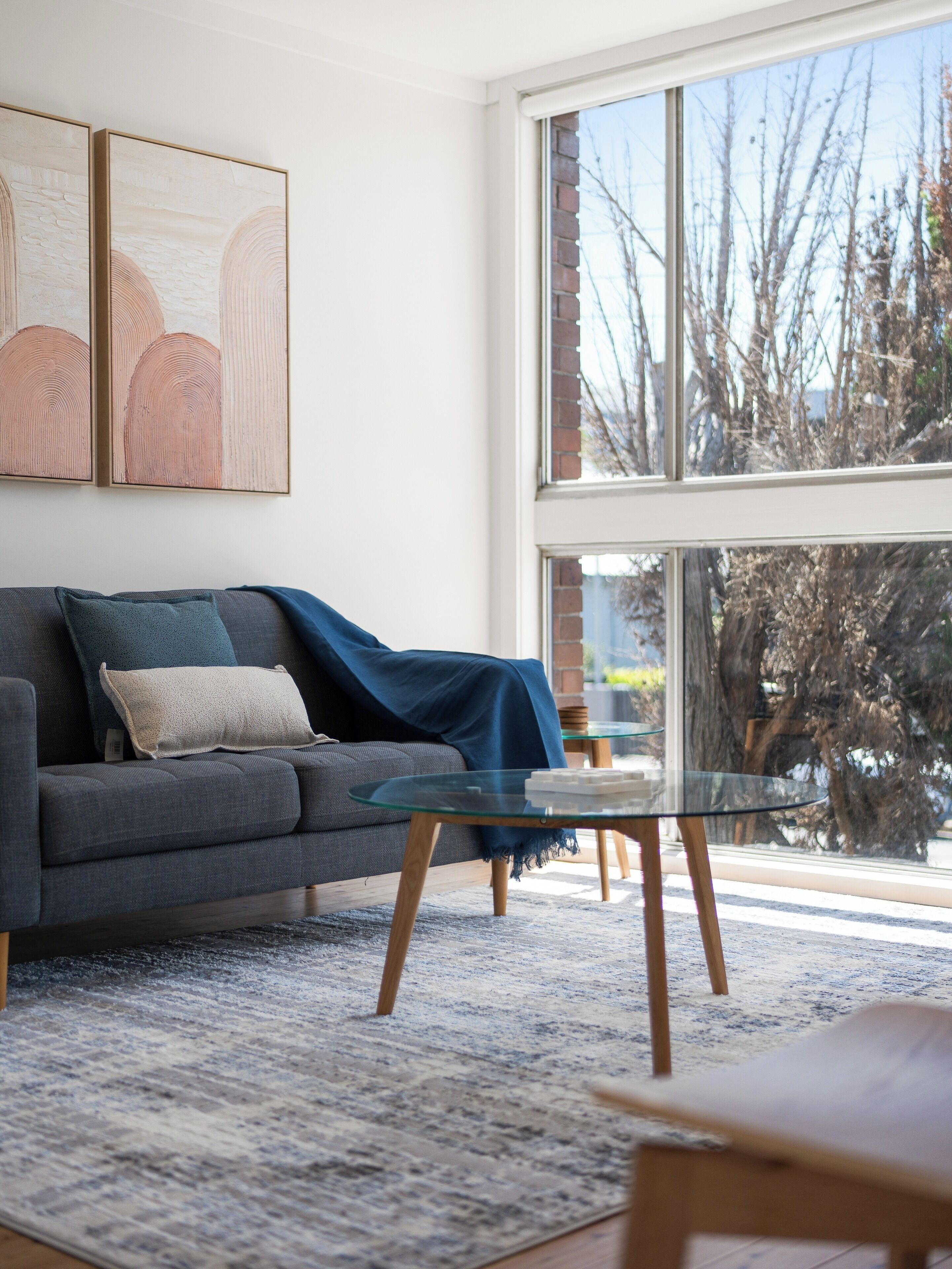 Una sala de estar moderna con un sofá gris, una mesa de centro de vidrio y un ventanal con vistas a árboles y un muro de ladrillo.