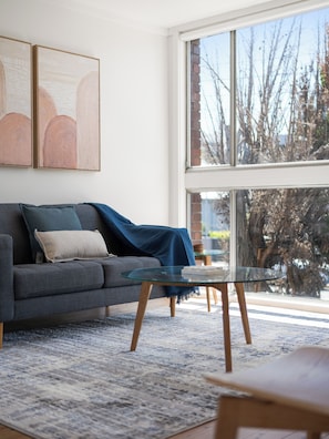 Una sala de estar moderna con un sofá gris, una mesa de centro de vidrio y un ventanal con vistas a árboles y un muro de ladrillo.
