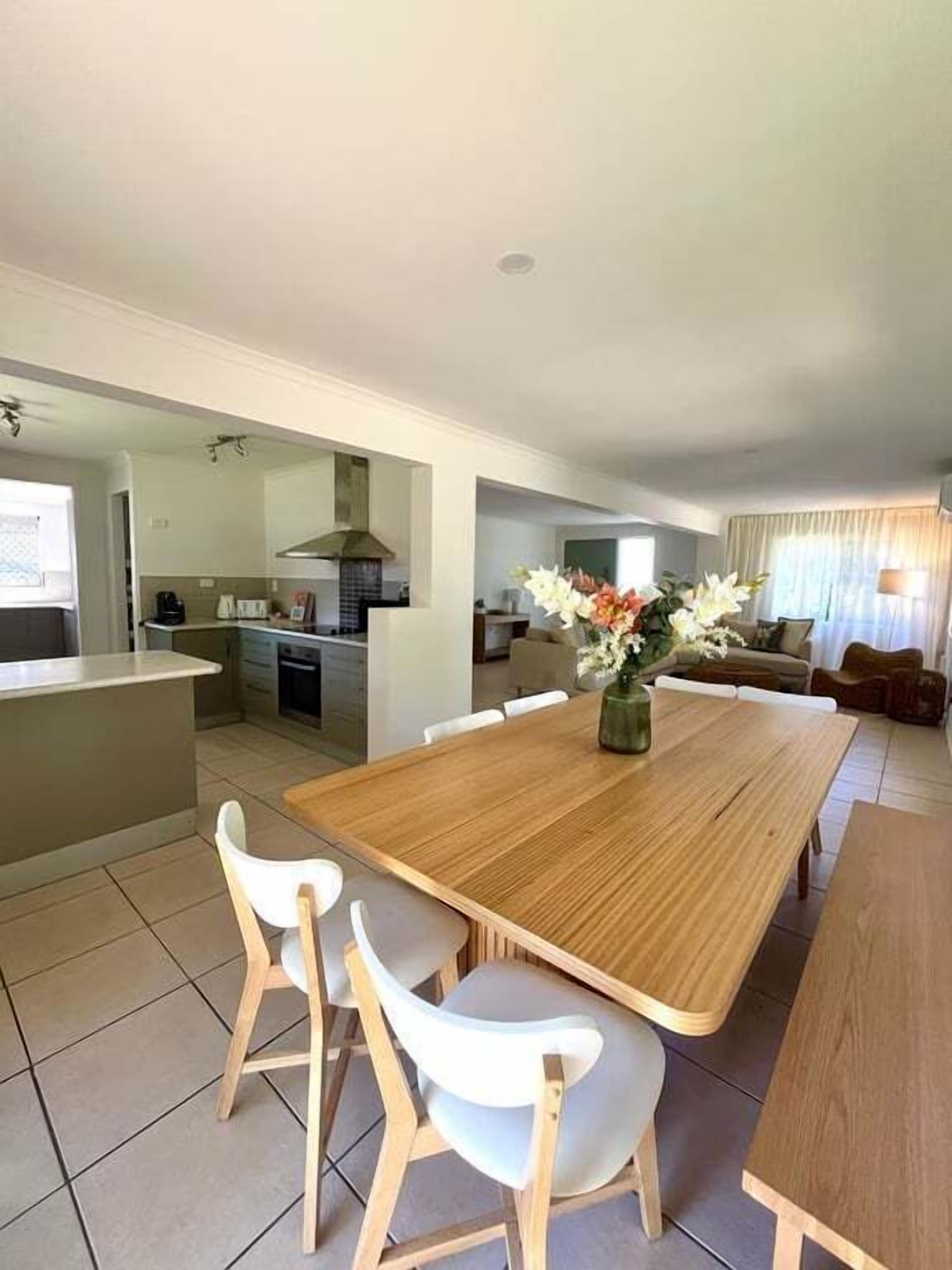 Une cuisine moderne dotée d’un îlot central, un coin repas avec une table en bois et des chaises blanches, et un salon avec un canapé et des rideaux.