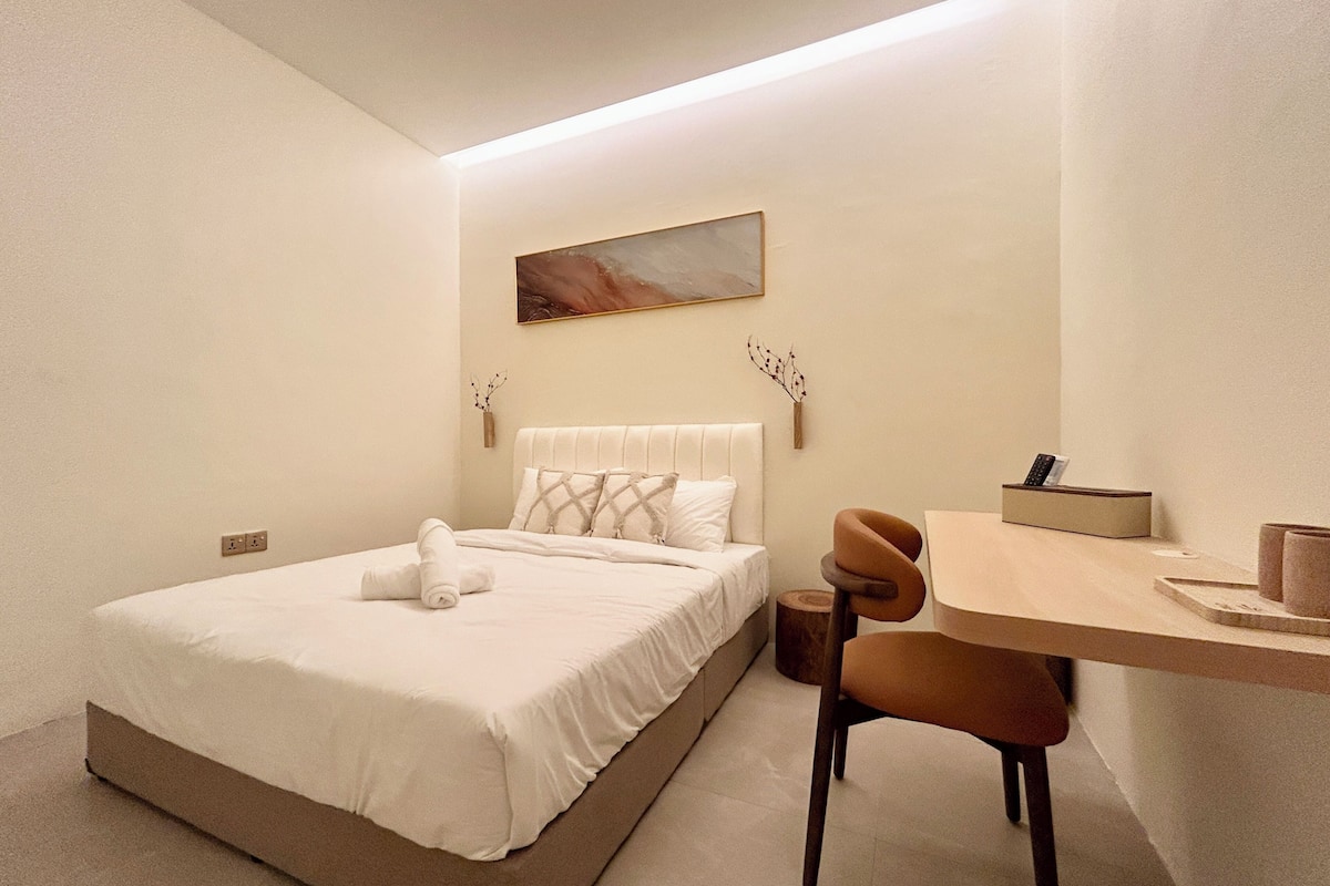 A bedroom with a bed, a desk, a chair, a painting on the wall, and a vase with branches.