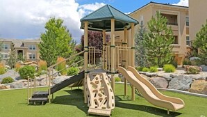 A playground with a slide and climbing structure, surrounded by a landscaped garden and residential buildings. - New Trendy 1BR Apartment with Heated Pool, Gym & Minigolf (Reno)