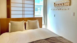 Hotellihuone, jossa on sänky, liukuva shoji-seinä, ilmastointilaite ja seinälle kiinnitetty pyykiteline.