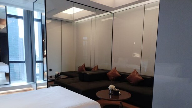 Une chambre d’hôtel moderne avec un grand lit, un canapé et une petite table. Un grand miroir orne l’un des murs, et une fenêtre offre une vue sur la ville.