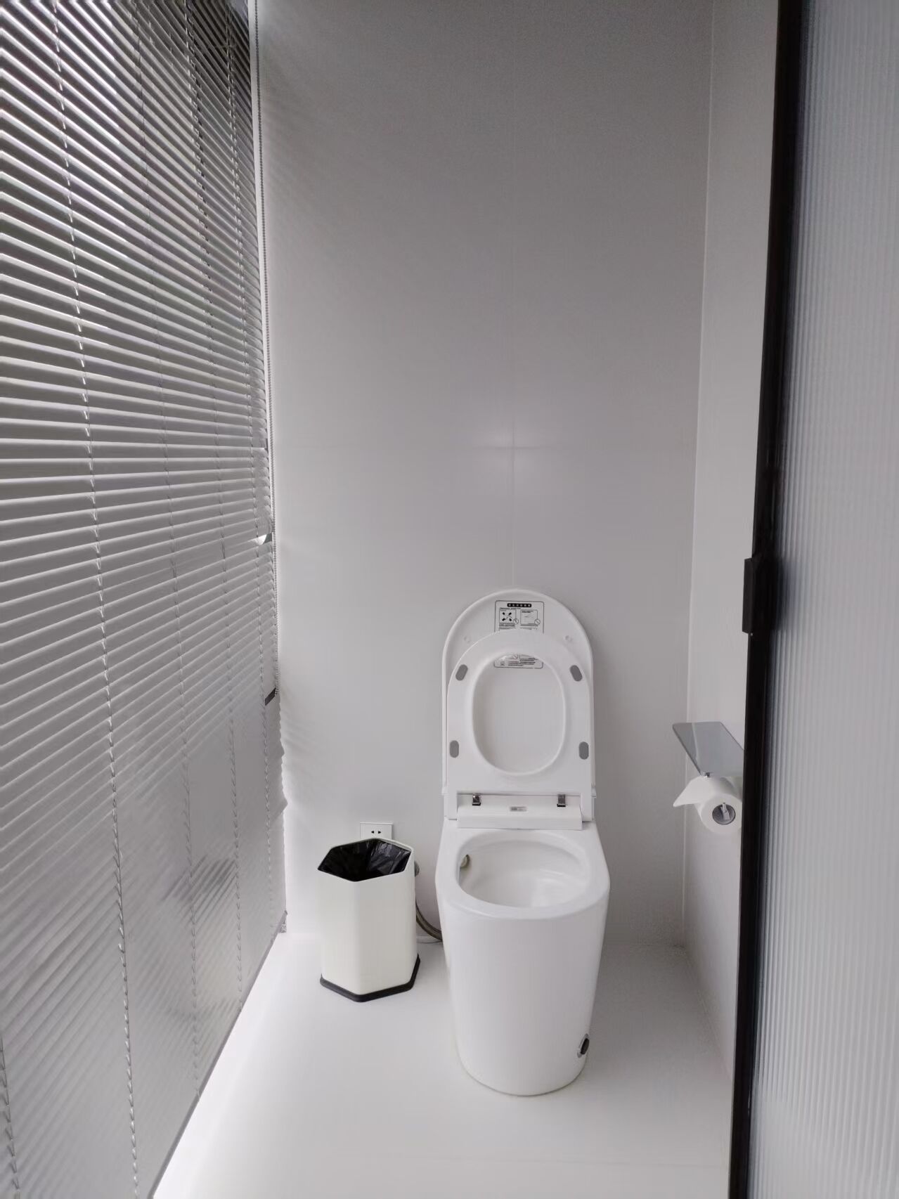 A small, white, modern bathroom with a toilet, a trash bin, and a window with blinds.