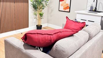 Un salon moderne avec un canapé gris, un coussin rouge, une plante en bambou et un coin cuisine doté d’un plan de travail blanc et d’un robinet noir.