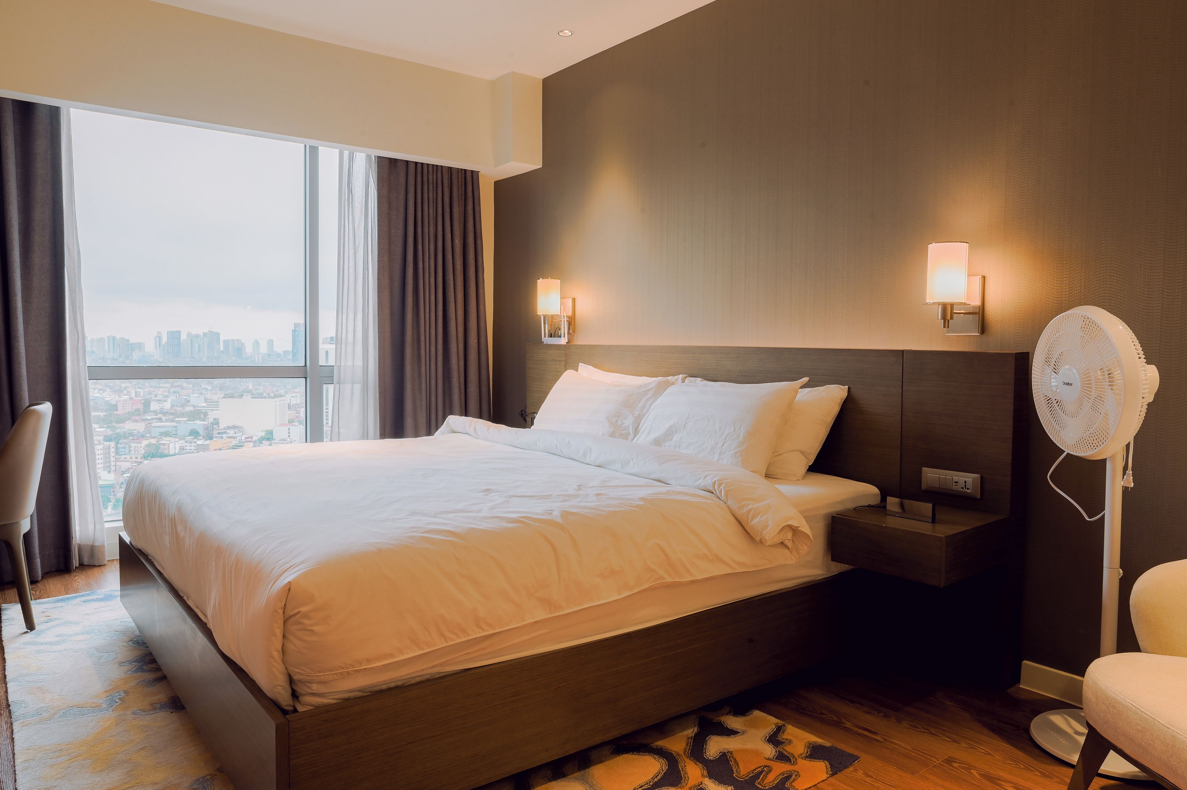 A modern hotel room with a large bed, two bedside lamps, a nightstand, a standing fan, and a view of the city through large windows.