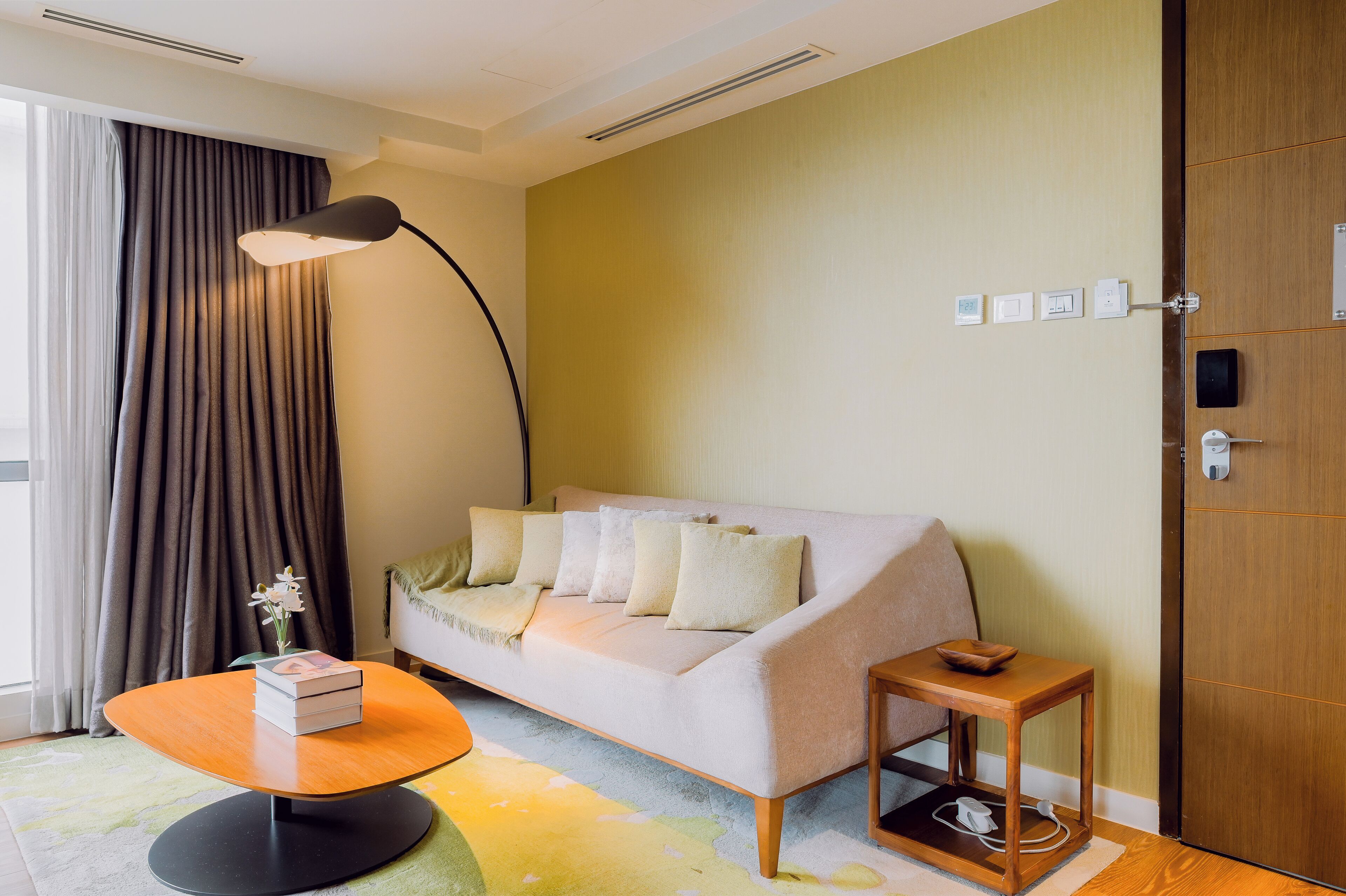A modern hotel room with a sofa, a small side table, a lamp, and a wooden door.
