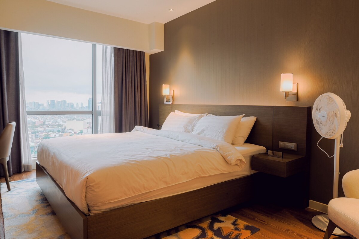 A modern hotel room with a large bed, two bedside lamps, a nightstand, a standing fan, and a view of the city through large windows.