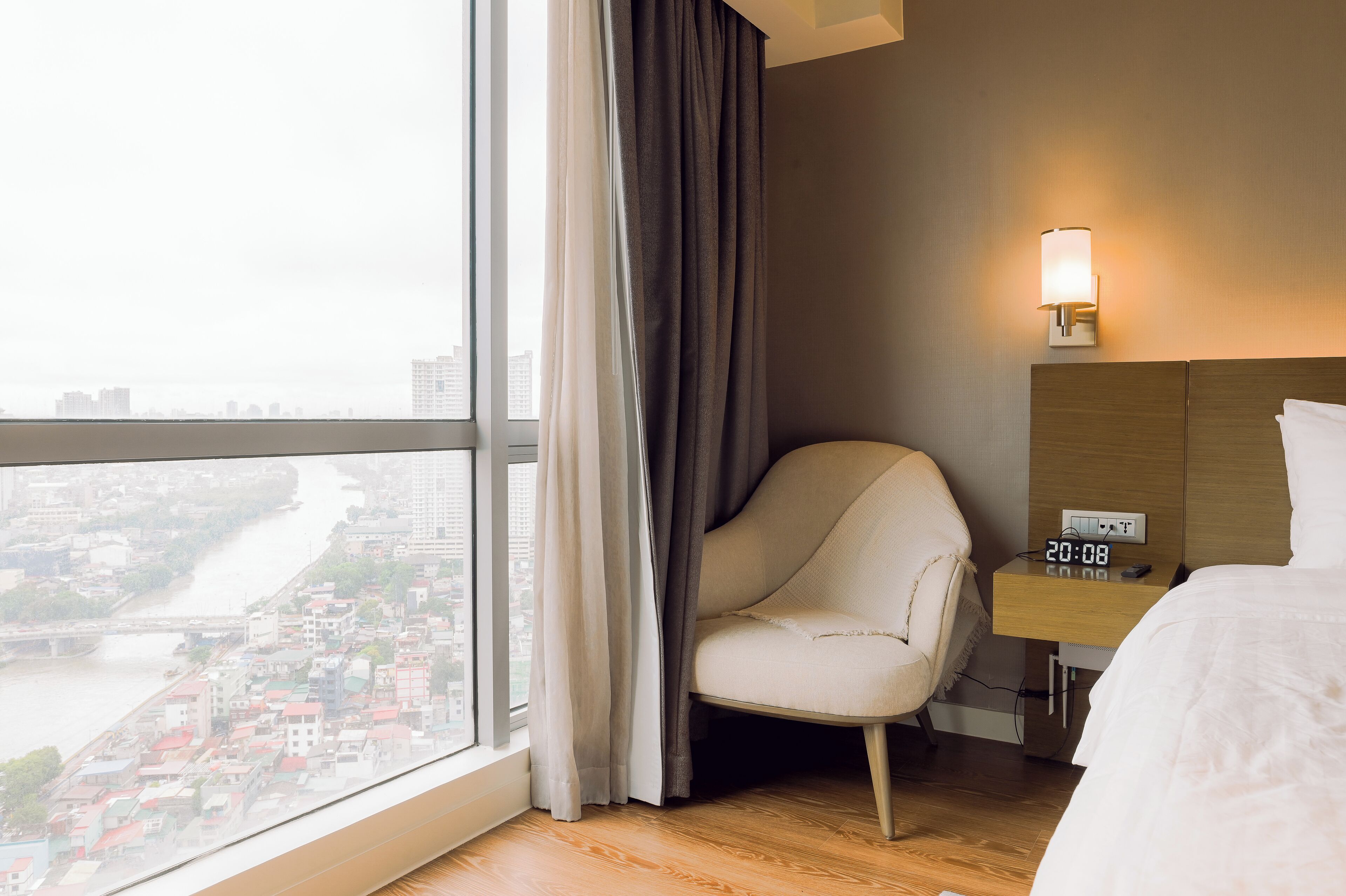 A hotel room with a large window, a chair, a bedside table with a clock, and a bed.