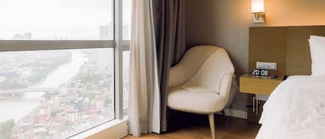 Habitación de hotel con un ventanal grande, una silla, una mesita de noche con un reloj y una cama.