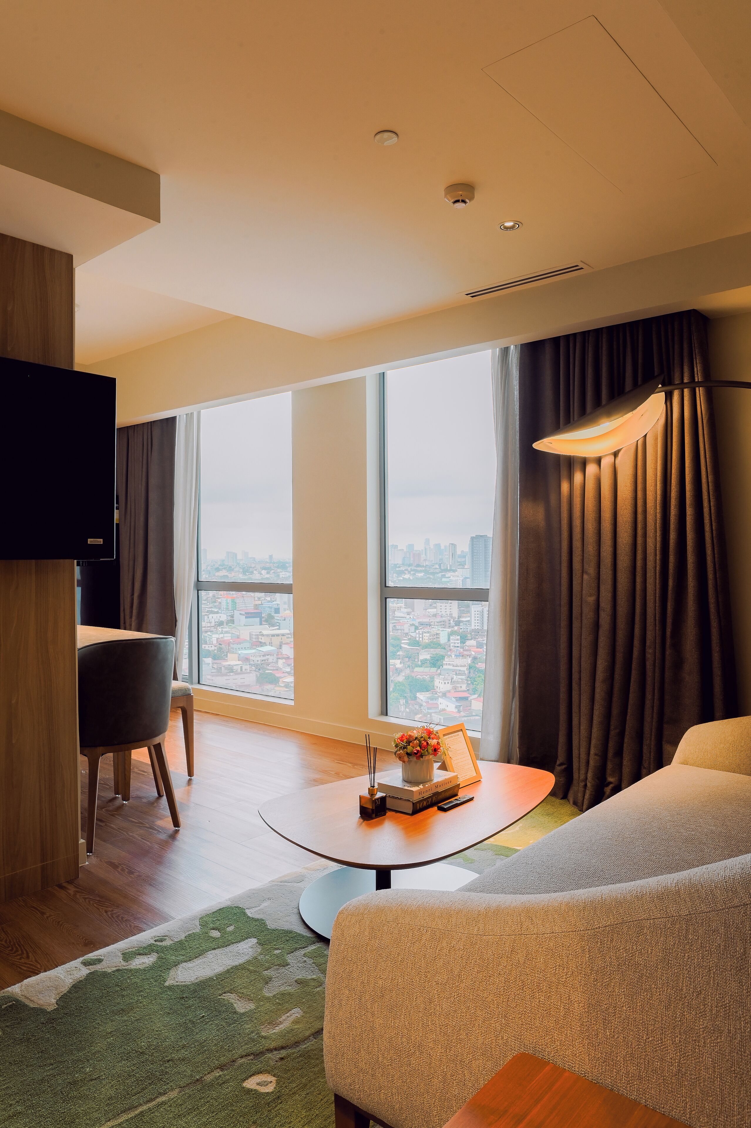 Une chambre d’hôtel moderne dotée d’une grande fenêtre, d’un téléviseur à écran plat, d’une table ronde, d’un canapé et d’un tapis vert.