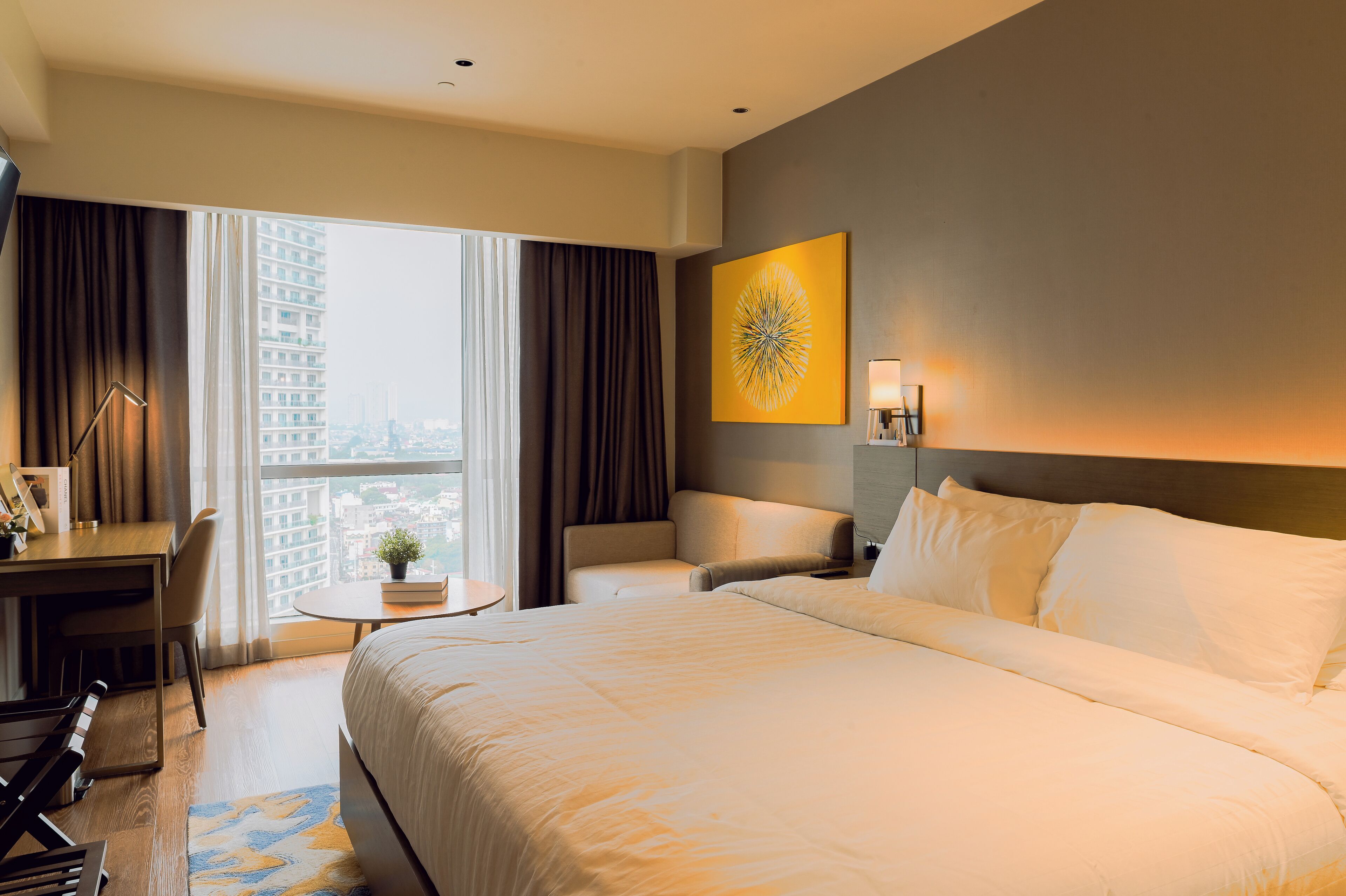 Une chambre d’hôtel avec un grand lit, un bureau, une chaise, une petite table et une vue sur la ville grâce à de grandes fenêtres.