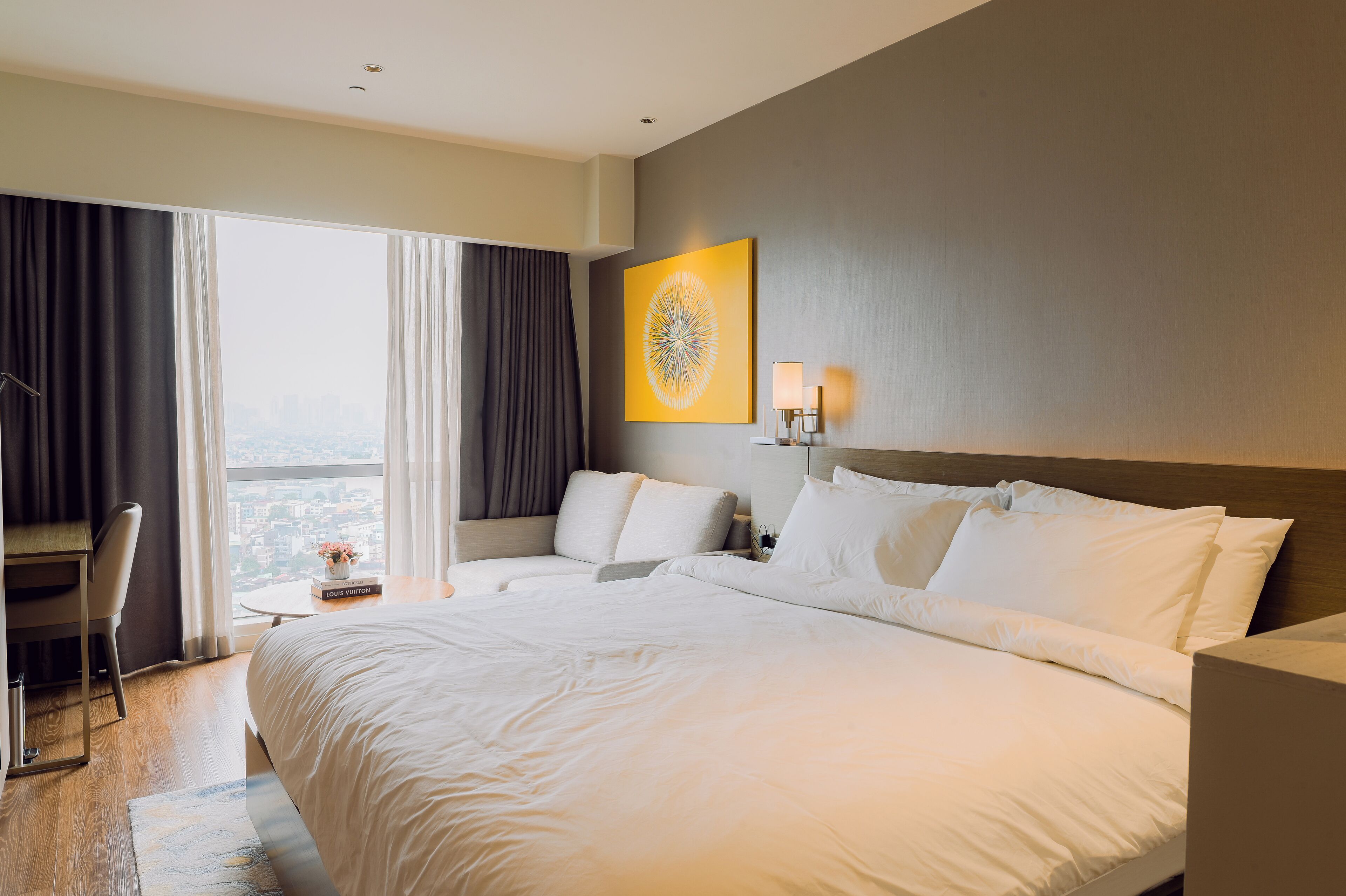 A hotel room with a large bed, a desk, a chair, and a view of the city through the window.
