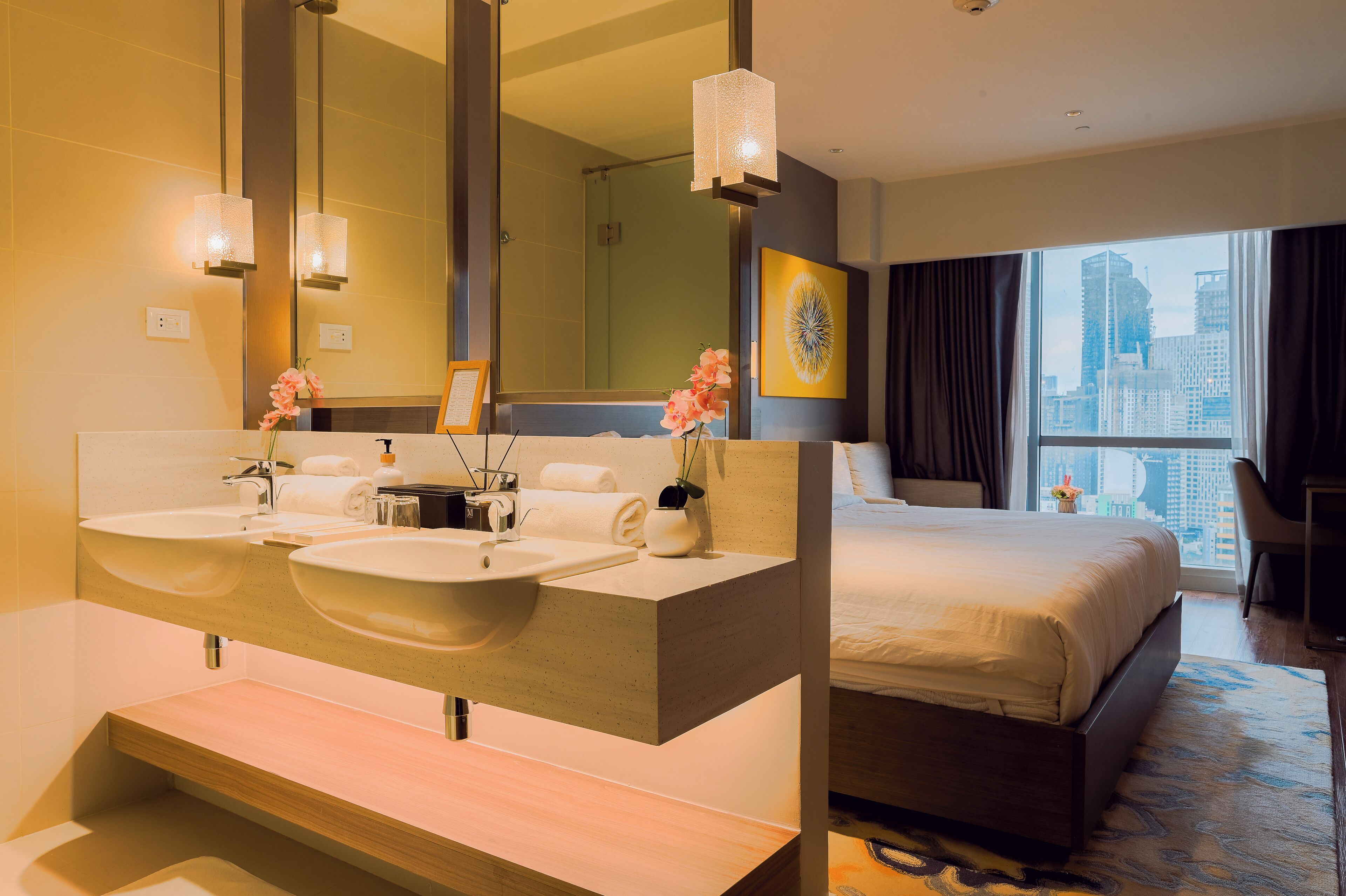 A hotel room with a large bed, a double sink vanity, and a city view through the window.