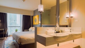A hotel room with a large bed, a double sink vanity, a large mirror, and a view of a cityscape through the window. - Hotel Residences at Acqua (Mandaluyong)