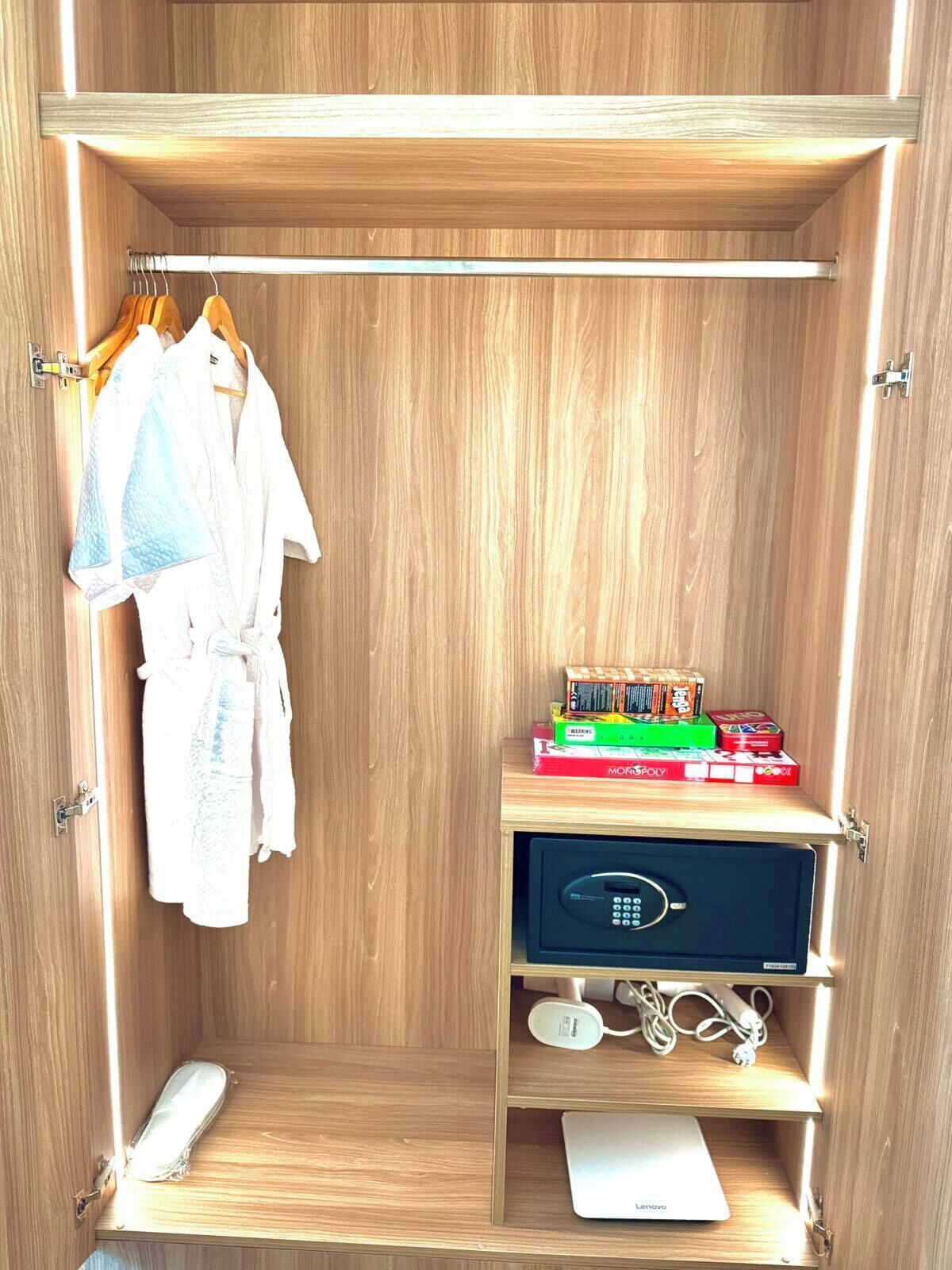 A wooden wardrobe with a hanging robe, a shelf with board games, a small electronic device, and a laptop.