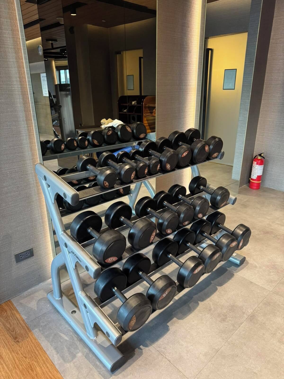 A gym with a mirrored wall, a fire extinguisher, and a rack of dumbbells.