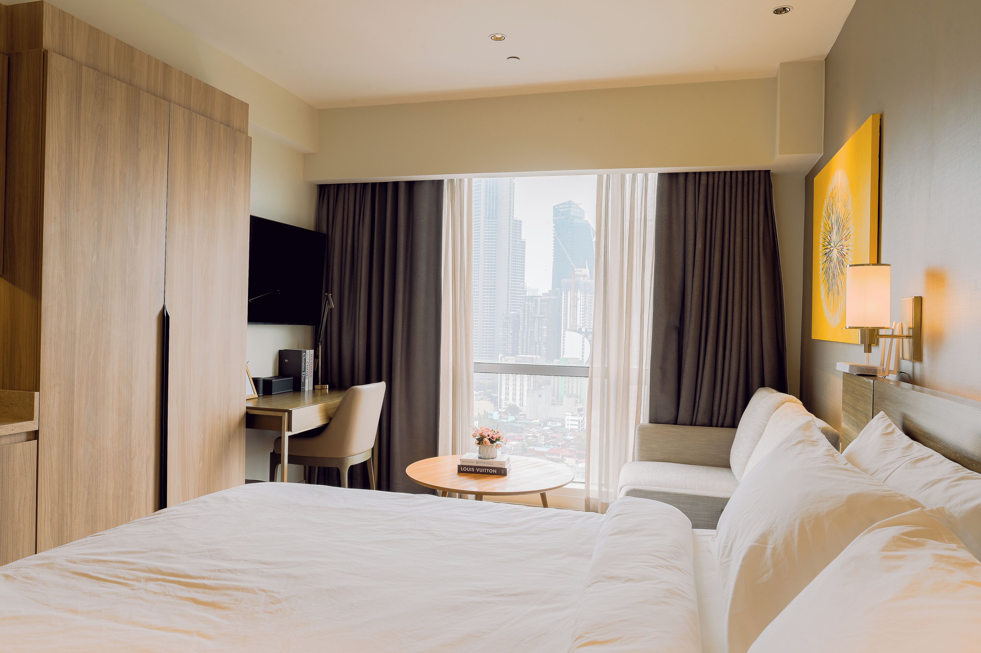 A hotel room with a large bed, a desk with a chair, a small table with a vase, and a view of the city through large windows.