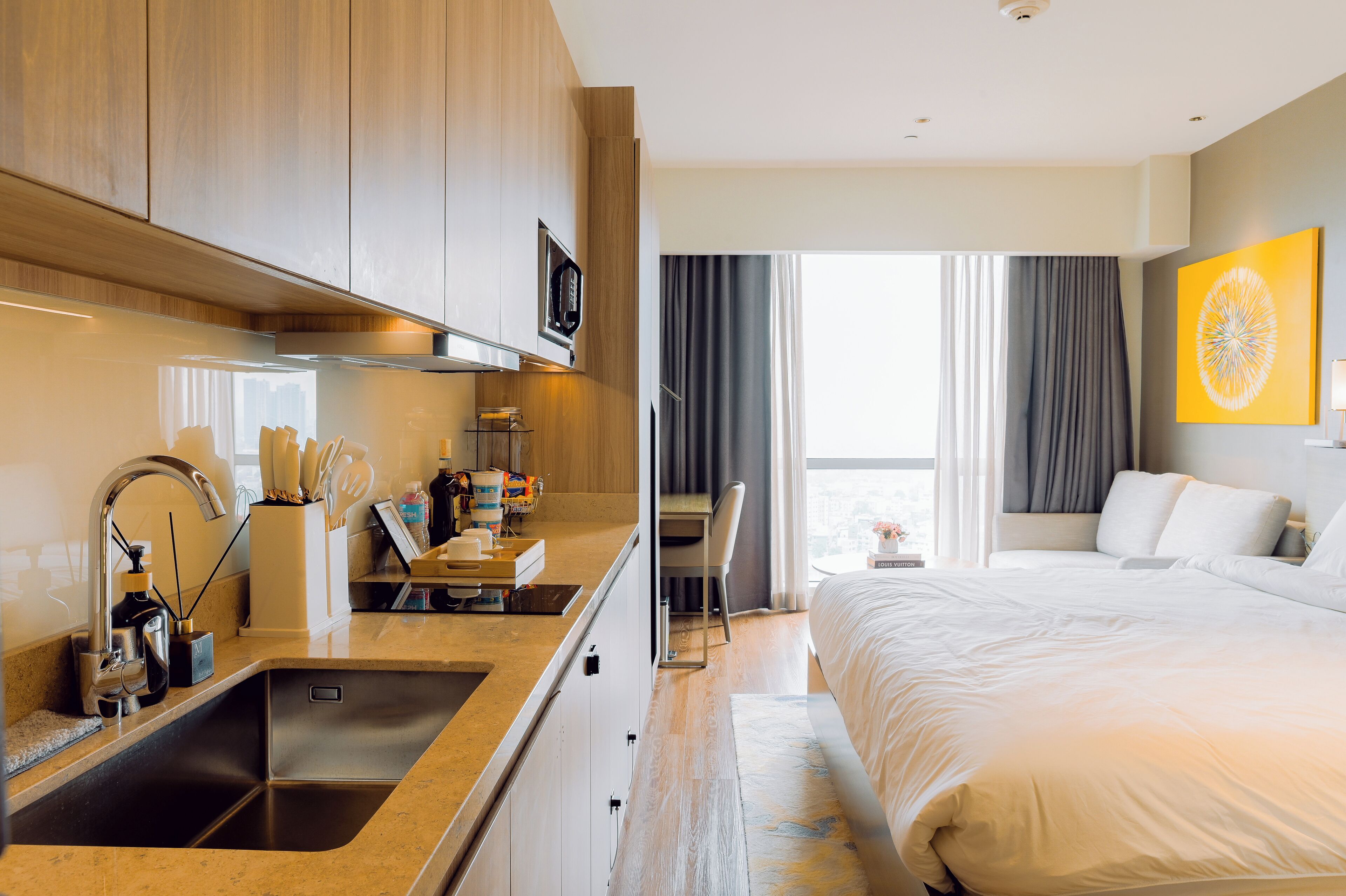 A modern hotel room with a kitchenette, a large bed, a desk, and a view of the city.