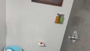 Un baño con pared de azulejos blancos, una ventana con persianas horizontales, un cubo azul sobre un taburete y una zona de ducha.