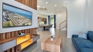 Ein modernes Wohnzimmer mit einem großen Fernseher, der eine Küstenstadt zeigt, einer Holz-Unterhaltungsecke, einem blauen Sofa und einem Essbereich mit Tisch und Stühlen.