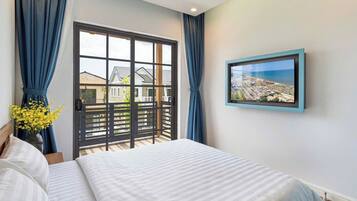A bedroom with a bed, a window with curtains, a view of buildings outside, and a framed picture on the wall.
