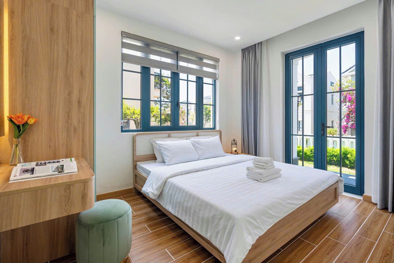 A modern hotel room with a large bed, a wooden desk, and a view of greenery outside.