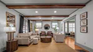 A spacious living room with a large sofa set, a coffee table, and a fireplace. There are two armchairs and a side table with a lamp. The room has wooden beams on the ceiling and large windows with curtains. - Hot Tub | Firepit | Putting Green | 5 Bedrooms (Shelbyville)
