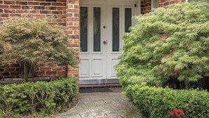 A brick house with a white front door, a small porch, and a well-maintained garden with various shrubs and a tree. - MetaWise Cozy Family Room4 in Shared Home Free Parking (Castle Hill)
