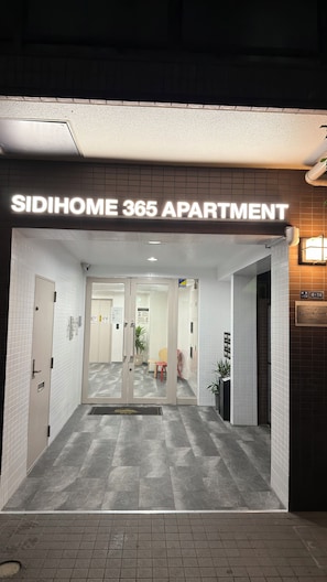 De ingang van SIDIHOME 365 APARTMENT, met een betegelde vloer, een bord boven de ingang en een deur aan de rechterkant.