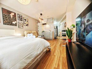 Quarto de hotel moderno com uma cama grande, televisão de tela plana, área de refeições e cozinha ao fundo.