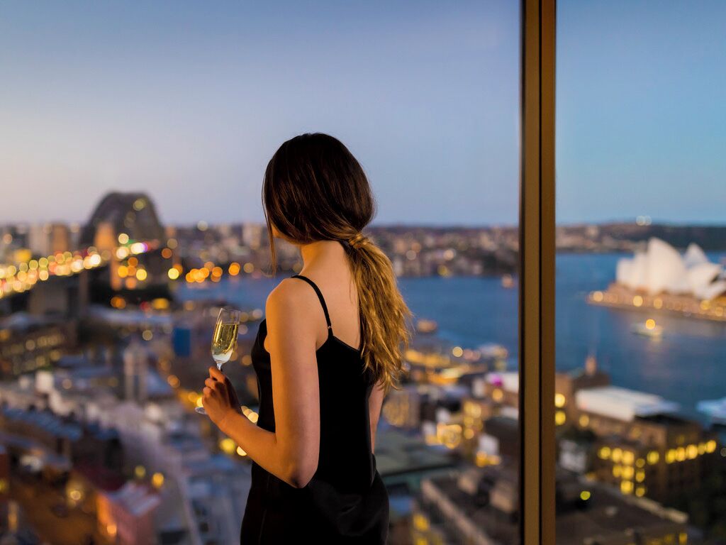 A person holding a glass of wine, overlooking a cityscape with a body of water and a prominent building in the distance.