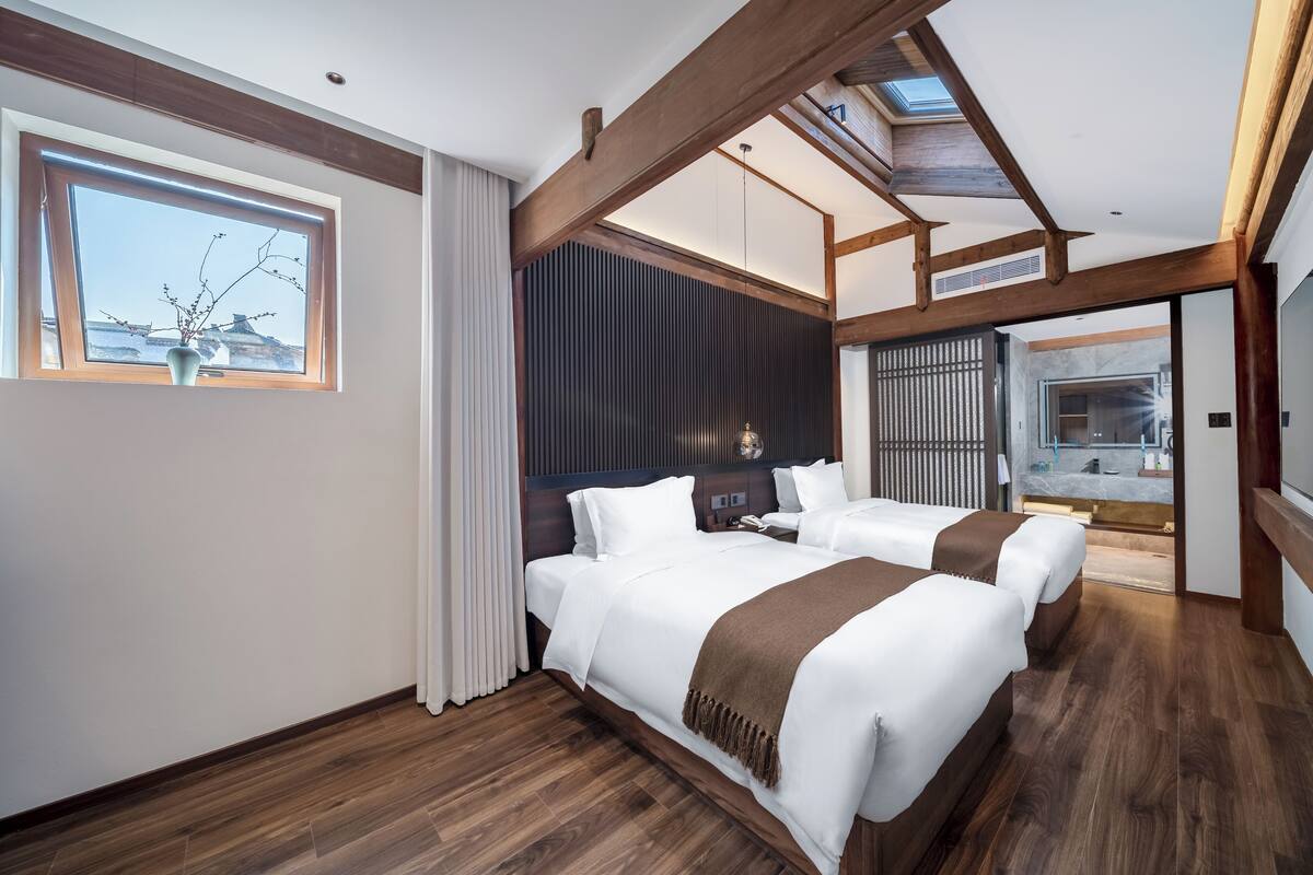 A modern hotel room with two beds, wooden flooring, a large window with a view, and a skylight.