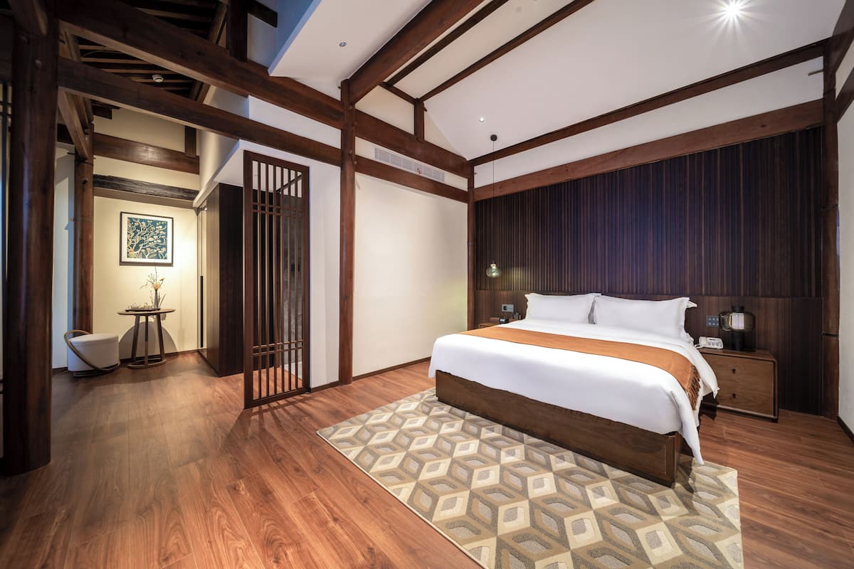 A modern bedroom with a large bed, wooden flooring, a patterned rug, and a nightstand with a lamp.