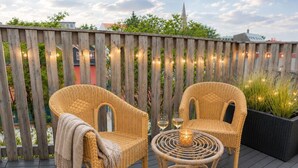 Una terraza de madera con mobiliario de mimbre, incluyendo dos sillas y una mesita con una vela y vasos.