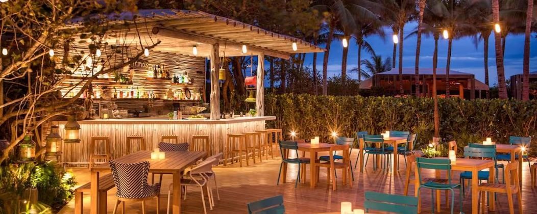 An outdoor bar area with wooden tables and chairs, surrounded by greenery and palm trees.