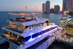 A large, multi-deck boat with a covered upper deck and a lower deck with seating areas, docked at a marina with city buildings in the background. - Casagrandesouthbeach by AmericanVacationLiving 4Rm (Miami Beach)