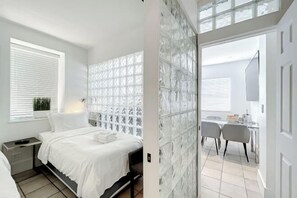 A modern bedroom with a bed, bedside table, and a glass partition wall. - Casagrandesouthbeach by AmericanVacationLiving 4Rm (Miami Beach)
